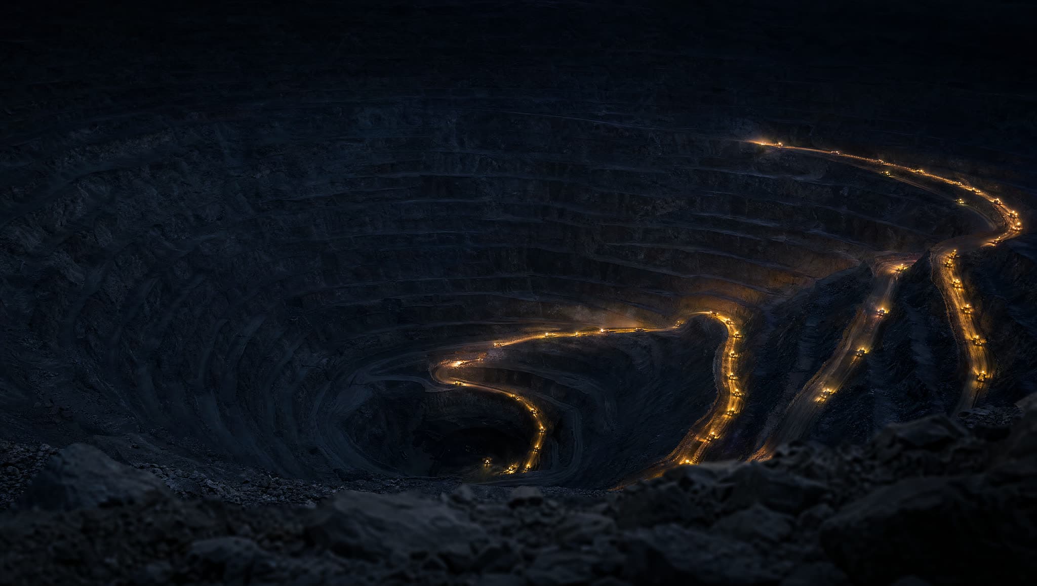 Tajo abierto de cordillera de noche con las luces doradas de camiones autónomos trazando las terrazas